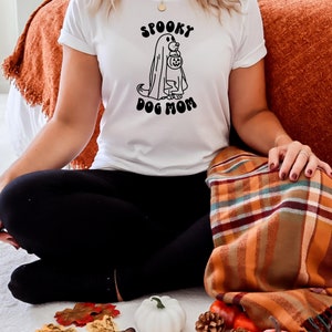 Op de afbeelding: Een wit T-shirt met een zwarte afbeelding van een hond die een spookkostuum draagt en een pompoen vasthoudt. De tekst "Spooky Dog Mom" staat onder de afbeelding.