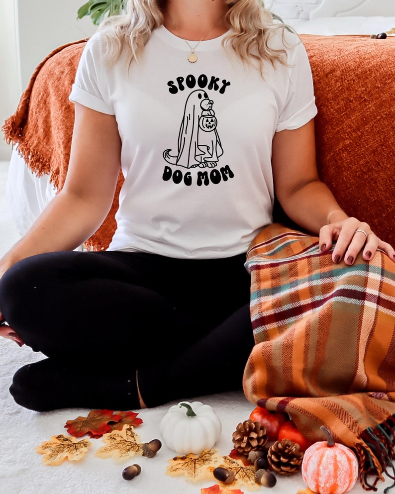 Op de afbeelding: Een wit T-shirt met een zwarte afbeelding van een hond die een spookkostuum draagt en een pompoen vasthoudt. De tekst "Spooky Dog Mom" staat onder de afbeelding.
