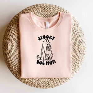 Op de afbeelding: Een perzikkleurig T-shirt met een zwarte afbeelding van een spookhond die een pompoen vasthoudt. De tekst "Spooky Dog Mom" staat onder de afbeelding.
