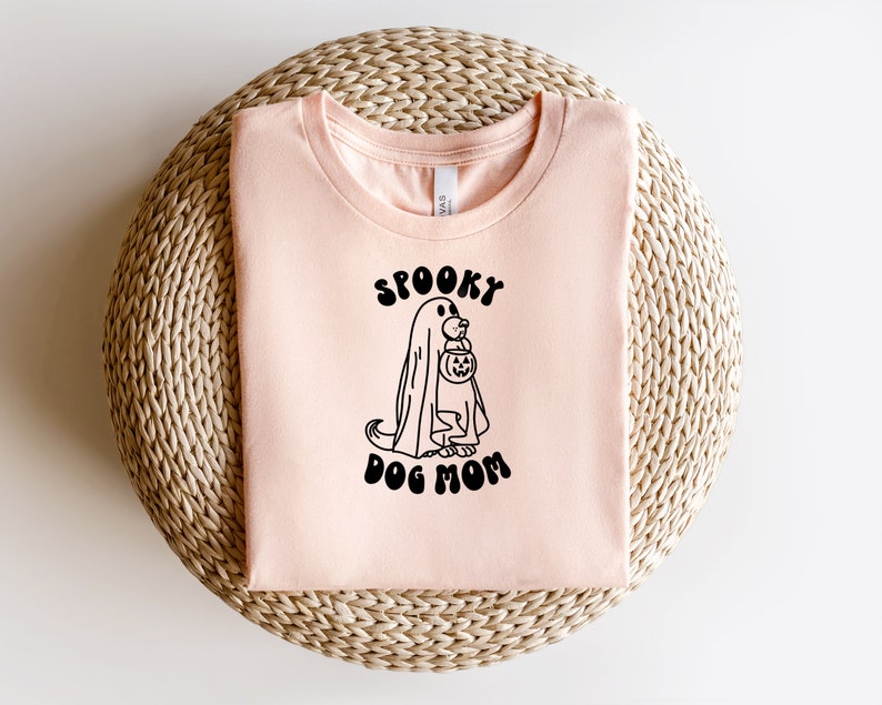 Op de afbeelding: Een perzikkleurig T-shirt met een zwarte afbeelding van een spookhond die een pompoen vasthoudt. De tekst "Spooky Dog Mom" staat onder de afbeelding.