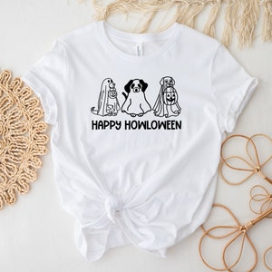 K&ouml;nnte beinhalten: Wei&szlig;es T-Shirt mit einer schwarz-wei&szlig;en Illustration von drei Hunden, die als Geister f&uuml;r Halloween verkleidet sind. Der Text "Happy Howloween" ist unter der Illustration gedruckt.