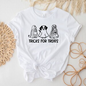 K&ouml;nnte beinhalten: Wei&szlig;es T-Shirt mit einer schwarz-wei&szlig;en Grafik von drei Hunden, die als Geister f&uuml;r Halloween verkleidet sind. Der Text "TRICKS FOR TREATS" ist unter der Grafik gedruckt.