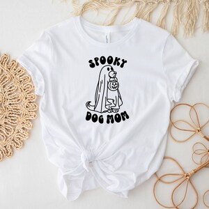 Op de afbeelding: Wit T-shirt met een zwart-witte illustratie van een hond die verkleed is als een geest en een pompoen vasthoudt. De tekst "Spooky Dog Mom" is in zwart onder de illustratie gedrukt.