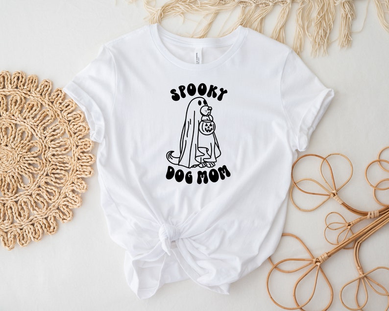 Op de afbeelding: Wit T-shirt met een zwart-witte illustratie van een hond die verkleed is als een geest en een pompoen vasthoudt. De tekst "Spooky Dog Mom" is in zwart onder de illustratie gedrukt.