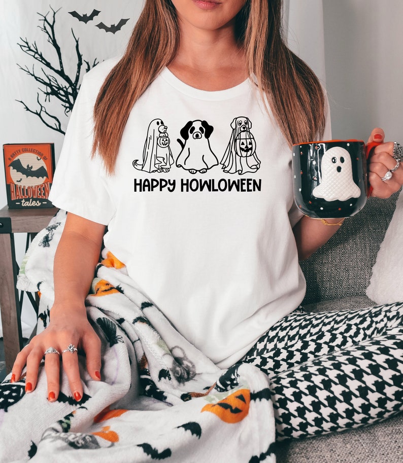 K&ouml;nnte beinhalten: Wei&szlig;es T-Shirt mit einer schwarzen Grafik von drei Hunden, die als Geister verkleidet sind und K&uuml;rbisse halten. Der Text "Happy Howloween" ist unter den Hunden gedruckt.