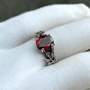May include: A silver ring featuring a large, oval-cut, deep red gemstone. The ring has an ornate, textured band with a detailed design. The gemstone is the focal point, set in a detailed, claw-like setting.