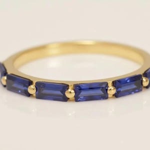 Op de afbeelding: Een gouden ring met rechthoekige, diepblauwe edelstenen. Het ontwerp van de ring omvat kleine gouden accenten tussen elke edelsteen, waardoor een doorlopend patroon ontstaat. De ring is geplaatst tegen een zachte, neutrale achtergrond.