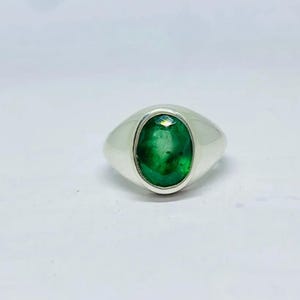 May include: A silver ring with a large, oval-cut emerald gemstone. The emerald is a vibrant green color and is set in a polished silver band. The ring is a classic design, suitable for everyday wear or special occasions.