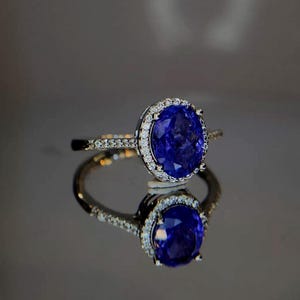 Può includere: Anello con una gemma ovale blu scuro, circondata da un'aureola di piccole pietre chiare. La fascia dell'anello è anch'essa ornata da piccole pietre chiare. L'anello è posto su una superficie riflettente, creando un effetto specchio.