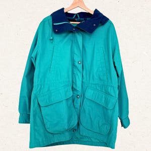 Può includere: Giacca color verde acqua con colletto e cappuccio blu navy, con ampie tasche frontali e chiusure con bottoni. La giacca è appesa a una gruccia di legno, che mostra uno stile vintage.