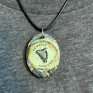 May include: A black cord necklace with a gold-colored pendant featuring a black and white image of a harp and the text "GUINNESS EXTRA STOUT"