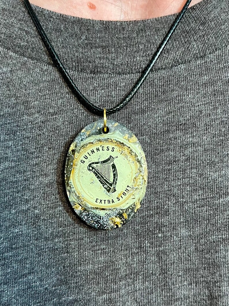 May include: A black cord necklace with a gold-colored pendant featuring a black and white image of a harp and the text "GUINNESS EXTRA STOUT"