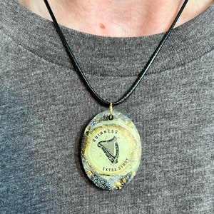 May include: A black cord necklace with a round pendant featuring a gold-colored Guinness logo with a harp and the text "GUINNESS EXTRA STOUT".