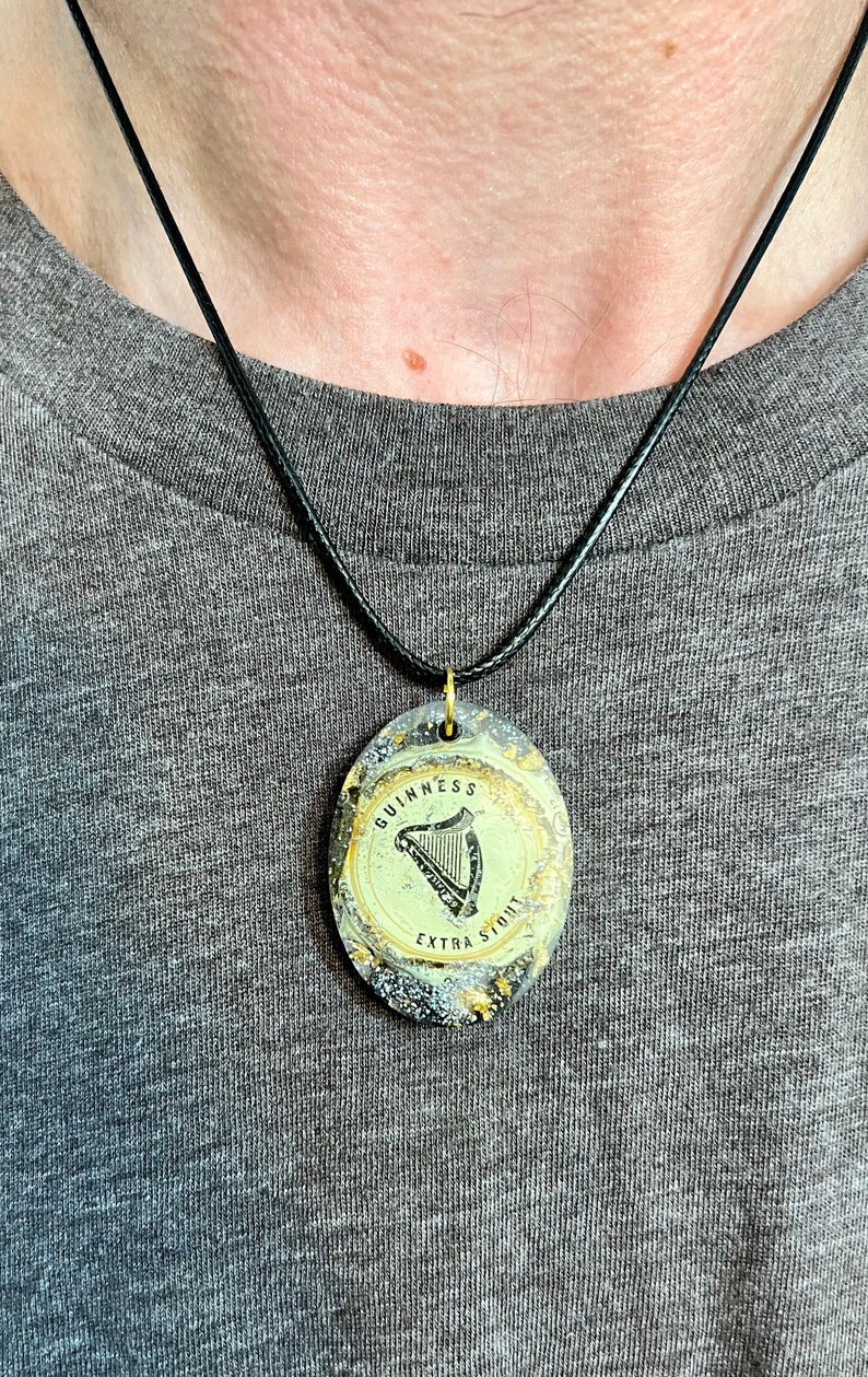 May include: A black cord necklace with a round pendant featuring a gold-colored Guinness logo with a harp and the text "GUINNESS EXTRA STOUT".