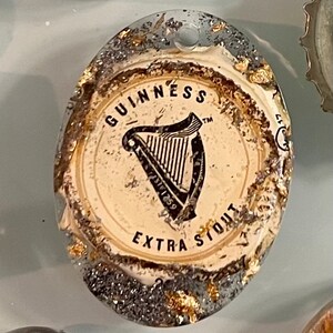 May include: A close-up of a round, white bottle cap with gold and silver glitter. The cap features the Guinness logo, a harp, and the text "GUINNESS EXTRA STOUT".