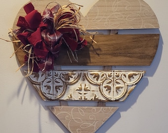 Könnte beinhalten: Eine herzförmige Wanddekoration aus Holz mit einem abgenutzten weißen und goldenen Fliesenmuster, einer braunen Holzplanke und einem beigefarbenen Hintergrund mit einem Wirbelmuster. Eine rote und braune Schleife mit einem Strohaccent ist an der Oberseite des Herzens befestigt.