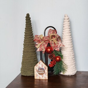 K&ouml;nnte beinhalten: Eine weihnachtliche Dekoration mit einer schwarzen Laterne, die mit roten Kugeln und einer karierten Schleife geschm&uuml;ckt ist. Zwei strukturierte, kegelf&ouml;rmige B&auml;ume, einer olivgr&uuml;n und einer cremefarben, flankieren die Laterne. Ein kleines hausf&ouml;rmiges Schild zeigt "Merry Christmas".