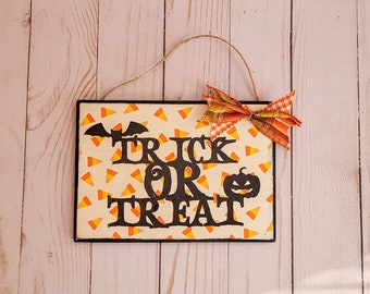 May include: A rectangular Halloween sign with a black border and a candy corn pattern background. The words "TRICK OR TREAT" are in black, with a bat and pumpkin graphic. A decorative bow is attached at the top.