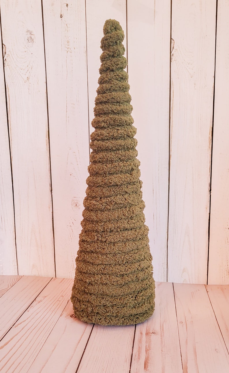 K&ouml;nnte beinhalten: Ein handgefertigter, olivgr&uuml;ner H&auml;kelbaum mit konischer Form. Der Baum besteht aus weichem, strukturiertem Garn und weist ein Spiraldesign auf. Der Baum ist ein dekorativer Artikel, der sich perfekt eignet, um jedem Raum einen Hauch von Natur zu verleihen.