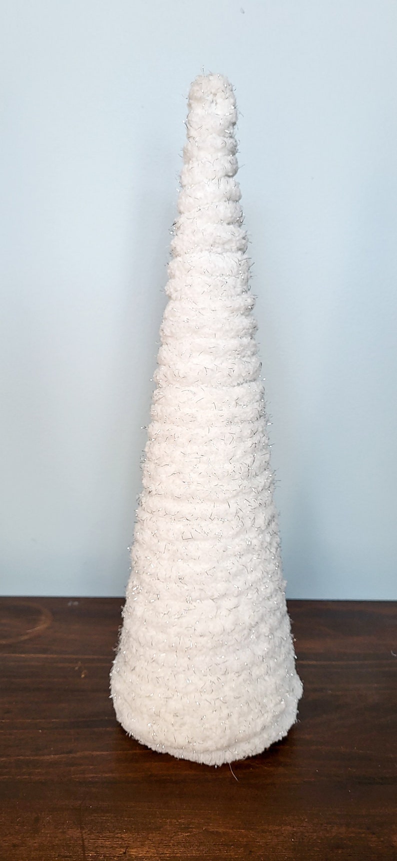 K&ouml;nnte beinhalten: Ein wei&szlig;er, kegelf&ouml;rmiger Dekobaum aus weichem, strukturiertem Material. Der Baum ist mit wei&szlig;en Fasern mit silbernen Glitzerakzenten bedeckt. Er steht auf einer dunkelbraunen Oberfl&auml;che vor einem hellblauen Hintergrund.