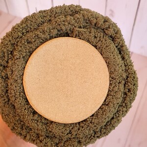 K&ouml;nnte beinhalten: Nahaufnahme einer Rolle olivgr&uuml;nen Chenille-Stoffs mit einem hellbraunen Holzsockel. Der Stoff hat einen strukturierten, geschlungenen Flor. Der Holzsockel ist kreisf&ouml;rmig und glatt. Der Stoff wird wahrscheinlich f&uuml;r Bastelarbeiten oder Heimdekoration verwendet.