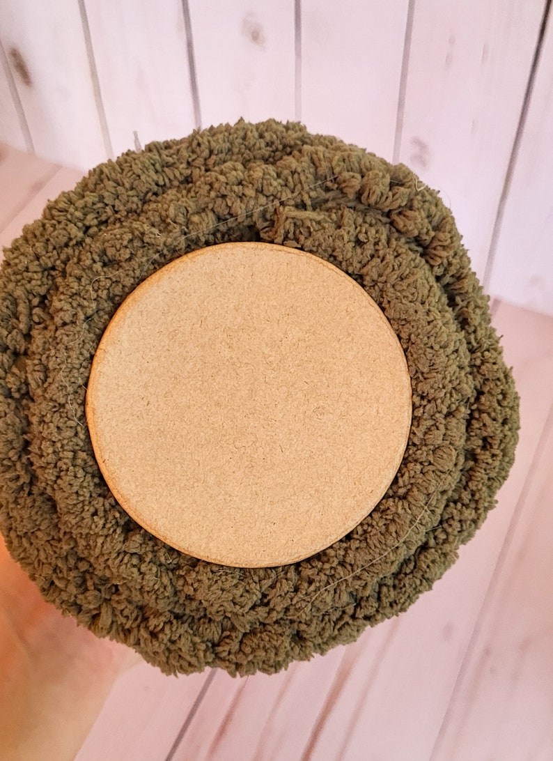 K&ouml;nnte beinhalten: Nahaufnahme einer Rolle olivgr&uuml;nen Chenille-Stoffs mit einem hellbraunen Holzsockel. Der Stoff hat einen strukturierten, geschlungenen Flor. Der Holzsockel ist kreisf&ouml;rmig und glatt. Der Stoff wird wahrscheinlich f&uuml;r Bastelarbeiten oder Heimdekoration verwendet.