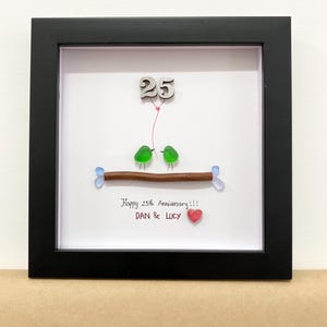 May include: A framed anniversary art piece with a black frame. The artwork features two green glass birds perched on a brown branch, with a red heart and the text "Happy 25th Anniversary!!! DAN & LUCY". A wooden number 25 is at the top.