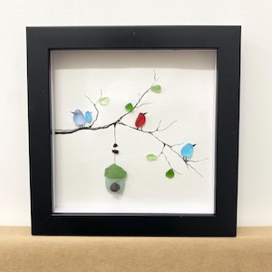 May include: Framed artwork depicting a branch with colourful birds and green leaves. The birds are blue and red, and the leaves are green. A green birdhouse hangs from the branch. The artwork is in a black frame.