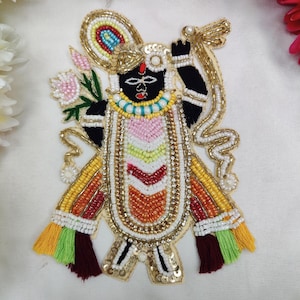 May include: A decorative embroidered patch featuring a stylized figure with a black face, adorned with intricate gold, white, pink, yellow, and red beadwork. The design includes a floral element, a curved gold border, and colorful tassels.