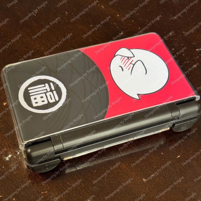 May include: A black and red Nintendo DS Lite case with a white cartoon cat design. The case has a clear plastic cover and a black base. The design includes a Japanese character in a white circle.