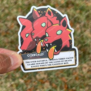 Puede incluir: Una pegatina con una ilustraci&oacute;n de dibujos animados de Cerbero, el perro de tres cabezas de la mitolog&iacute;a griega. Las cabezas de perro rojas tienen detalles amarillos y ojos verdes. El texto dice "Cerberus, Hound of Hell".