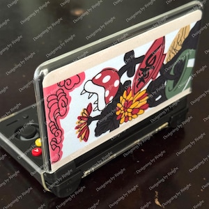 May include: A black handheld gaming console with a decorative cover featuring a colorful design. The cover includes a red and white mushroom, a yellow and red flower, and other graphic elements. The console has red, yellow, and black buttons.