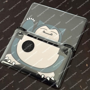 May include: Clear protective case for a gaming console featuring a cartoon Snorlax design in blue, white, and gray. The case is designed to fit the console and includes cutouts for buttons and vents. The design is repeated on the top cover.