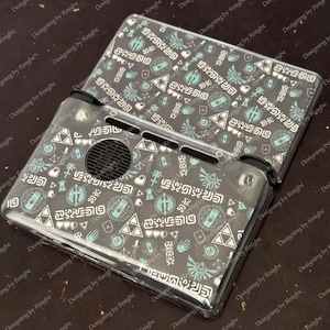 May include: A clear protective case for a handheld gaming device. The case has a repeating pattern of teal, white, and black symbols and text. The device is open, showing the screen and controls.