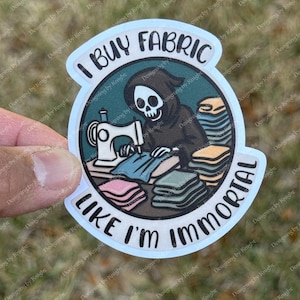 May include: A sticker with a cartoon grim reaper sewing fabric on a white sewing machine. The sticker has the text "I BUY FABRIC LIKE I'M IMMORTAL" in a circular design with a light blue border. Stacks of fabric are in the foreground.
