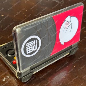 May include: A black and red Nintendo 3DS console with a custom cover. The cover features a white graphic and Japanese text within a white circle on a black background. The other side has a white cartoon character on a red background.