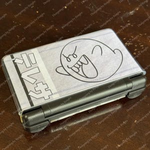Może przedstawiać: Czarna konsola do gier przenośna Nintendo 3DS z przezroczystą osłoną z kreskówkowym wzorem ducha Boo. Osłona ma również japoński tekst i szaro-białe paski w tle.