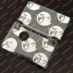 May include: Clear protective case for a handheld gaming device, featuring a repeating pattern of white cartoon ghosts on a gray background. The case is designed to fit the device snugly, with cutouts for buttons and speakers.
