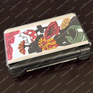 May include: A black handheld gaming console with a clear protective cover featuring a colorful design. The design includes a red mushroom, flowers, and Japanese-style art. The console is rectangular and has a screen and buttons.
