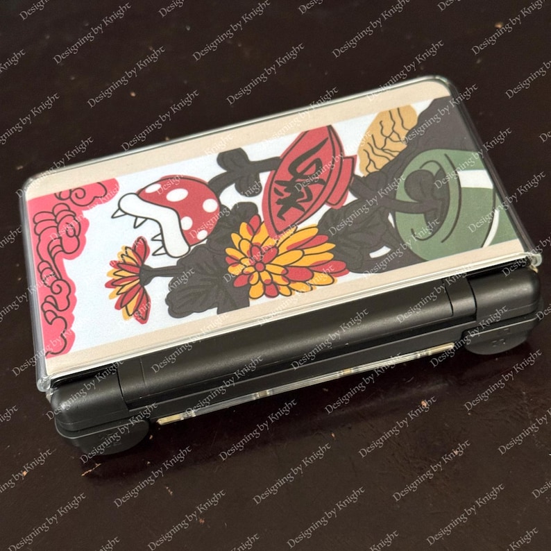 May include: A black handheld gaming console with a clear protective cover featuring a colorful design. The design includes a red mushroom, flowers, and Japanese-style art. The console is rectangular and has a screen and buttons.