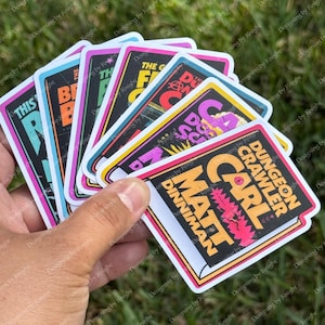 May include: A hand holding a fan of colorful stickers. Each sticker features a unique design with text, including "DUNGEON CRAWLER CARL" and "MATT DINNIMAN" in bold fonts, set against vibrant backgrounds of black, pink, and blue.