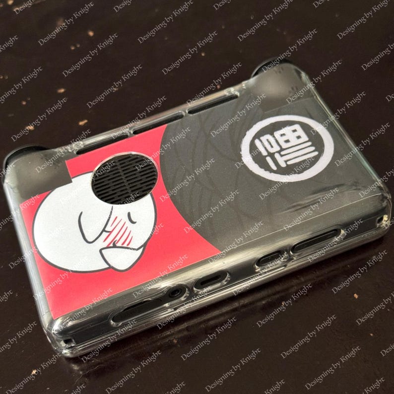 May include: Clear protective case for a handheld gaming device. The case features a red and black design with a white cartoon character and the text "10日日" in a white circle. The case is made of clear plastic.