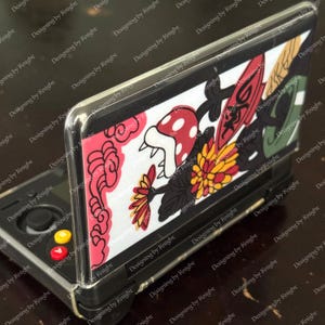 May include: A black handheld gaming console with a clear protective case featuring a colorful design. The design includes a red mushroom, flowers, and other graphic elements on a white background. The console has red, yellow, and black buttons.