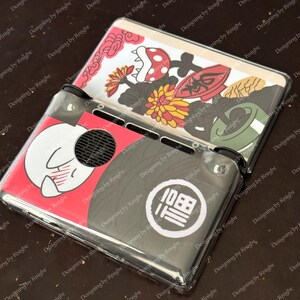 May include: A clear plastic Nintendo Switch case with colorful Japanese-inspired graphics. The case features a red and white design with a cartoon character, and a black and gray design with a white circular emblem.
