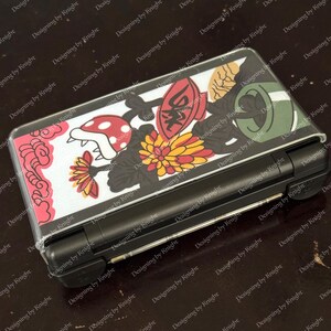 May include: A black handheld gaming console with a clear protective cover featuring a colorful design. The cover displays a red mushroom, yellow and red flowers, and other graphic elements. The console is closed and resting on a dark surface.