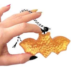 May include: A gold-colored bat-shaped ornament with a textured surface. The bat is suspended by a black and white cord. The ornament is held up by a hand with manicured nails.