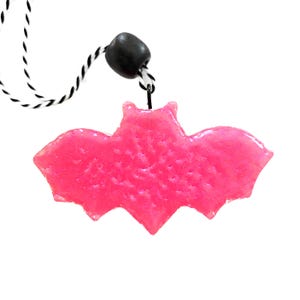 May include: A bright pink, glittery bat-shaped pendant suspended from a black and white twisted cord. The bat figure features a textured surface and is attached to the cord by a small black bead, creating a distinct contrast between the vibrant pink and the monochrome string.