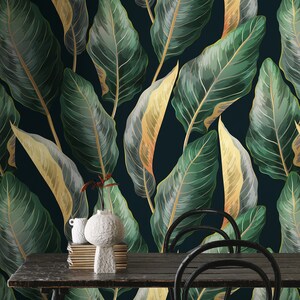 Peut inclure: Un fond bleu fonc&eacute; avec un motif r&eacute;p&eacute;titif de grandes feuilles vertes et dor&eacute;es. Une table en bois avec deux chaises noires se trouve devant le fond &agrave; motifs. Un vase en c&eacute;ramique blanc avec une pile de livres est pos&eacute; sur la table.