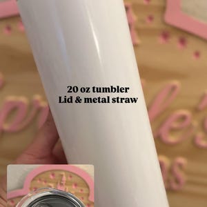May include: A white 20 oz tumbler with a smooth finish, featuring a clear plastic lid and a metal straw. The text 20 oz tumbler Lid & metal straw is printed on the side of the cylindrical container, which has a flat base and a circular opening.