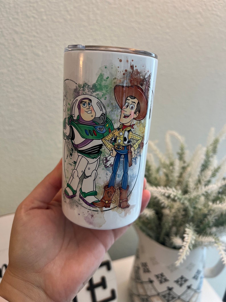 May include: A white cylindrical tumbler featuring a colorful watercolor-style graphic of the characters Buzz Lightyear and Woody from Toy Story. The design includes splashes of green, blue, and brown paint, and the tumbler has a clear plastic lid.